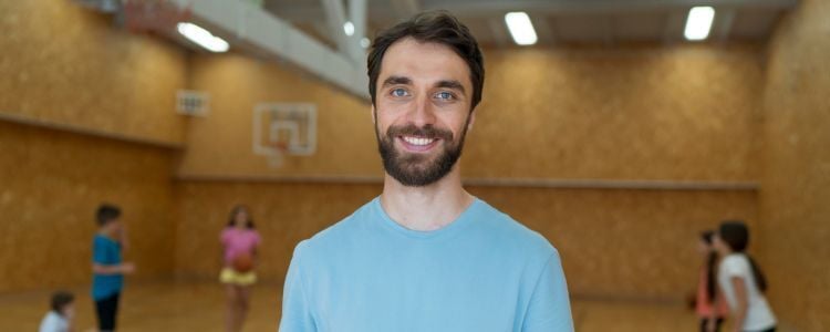 Profesor de educación física sostiene una tabla con clip y hojas mientras supervisa a un grupo de niños que se pasan un balón de básquetbol