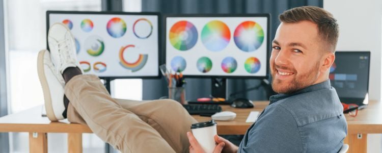 Diseñador gráfico digital sonríe sentado cómodamente con un vaso de café frente a su espacio de trabajo, que incluye un escritorio con tres pantallas donde se aprecian gráficos de colores