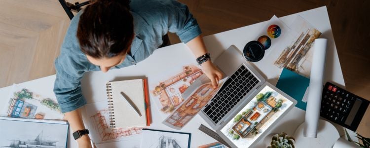 Diseñadora gráfica digital trabaja sobre escritorio con diferentes bocetos impresos que también aparecen en la pantalla de una laptop y una tablet en que realiza selección de colores, entre otras herramientas como calculadora, lámparas y libreta