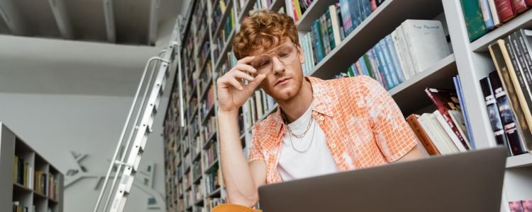 Hombre joven trabaja entre los anaqueles de una biblioteca mientras analiza contenido en su laptop