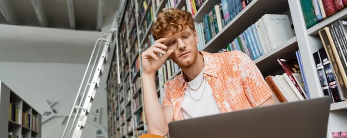 Hombre joven trabaja entre los anaqueles de una biblioteca mientras analiza contenido en su laptop
